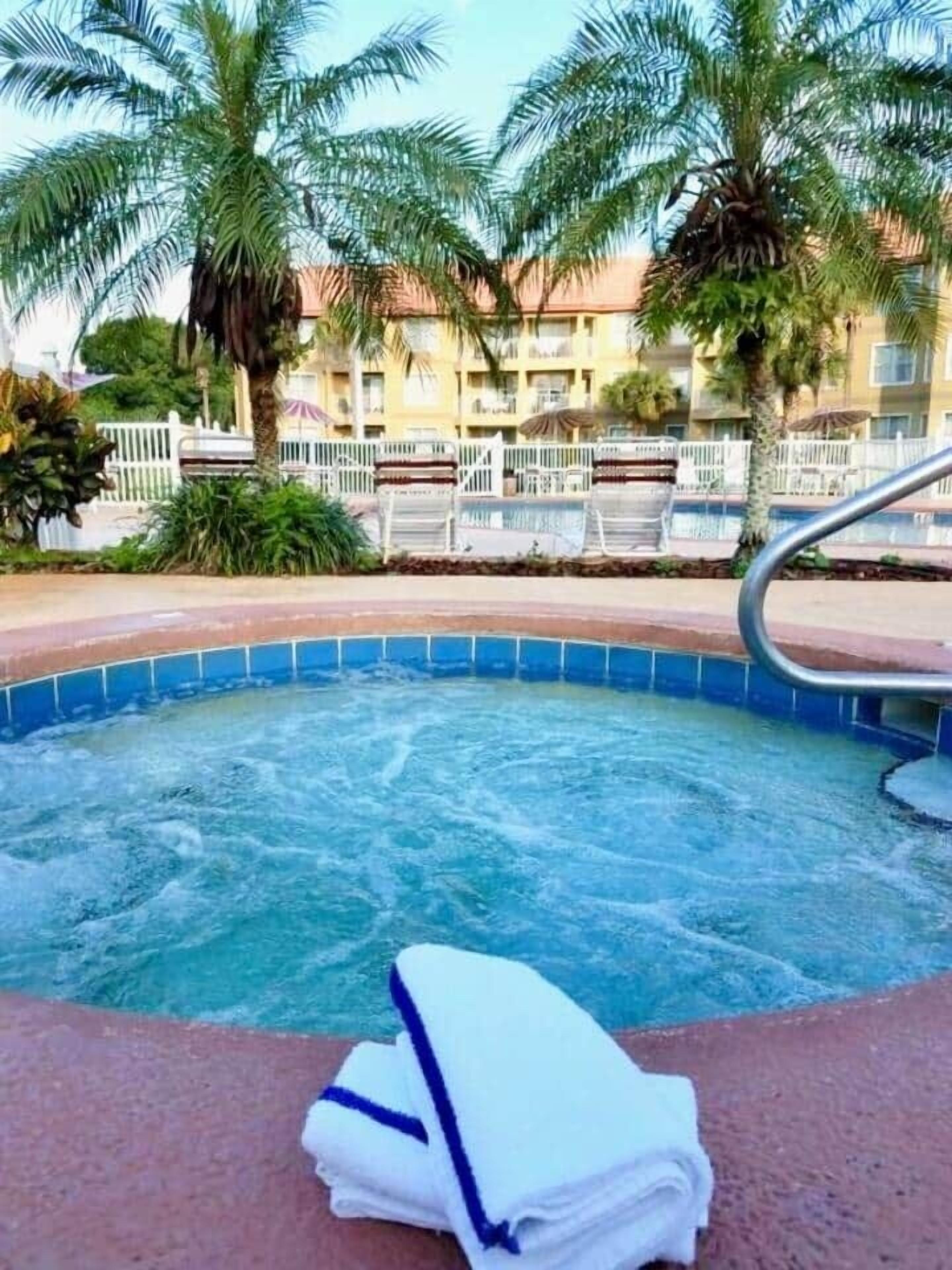 A hot tub with a blue edge, surrounded by palm trees and a poolside area with lounge chairs.