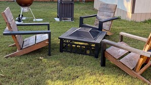 Un jardin avec une terrasse en bois, un barbecue, un couvercle pour barbecue, une poubelle et deux chaises en bois.