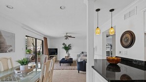 Ein modernes Wohnzimmer mit einem Glastisch, einem Deckenventilator und einer Küchenzeile mit schwarzer Arbeitsplatte und einer Uhr.