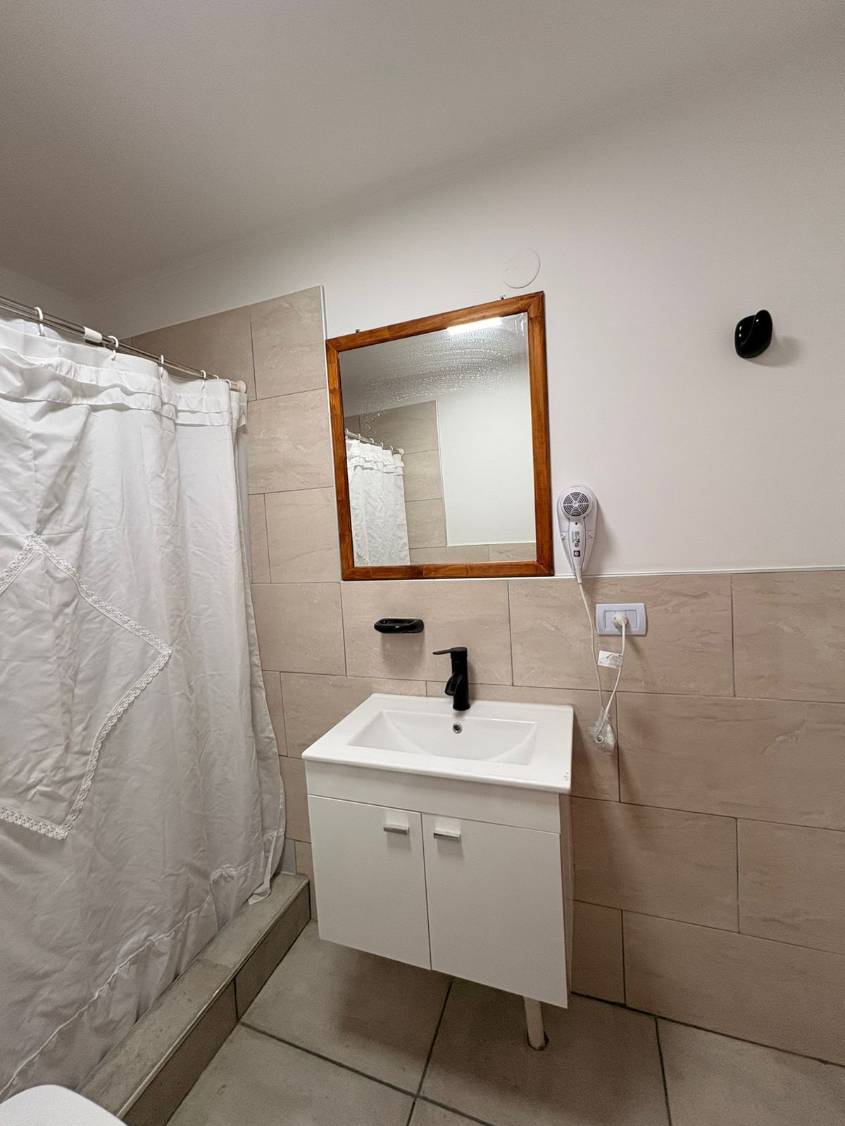 A bathroom with a white sink, a mirror, a white shower curtain, and a hairdryer on the wall.