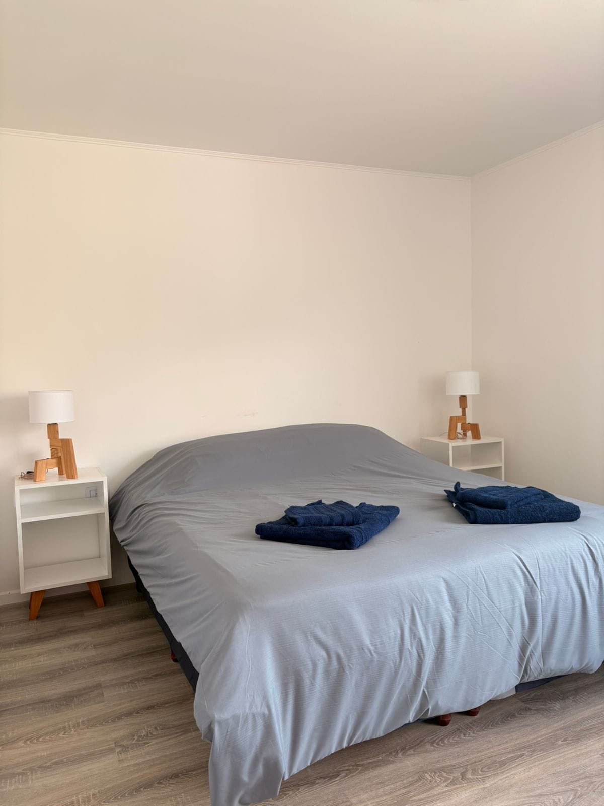 A neatly made bed with a grey bedspread and two folded blue towels, flanked by white nightstands with wooden lamps.