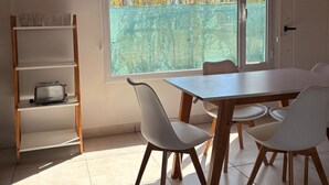 Un coin repas avec une table et des chaises, une étagère contenant des ustensiles de cuisine et une fenêtre donnant sur des arbres et une maison.