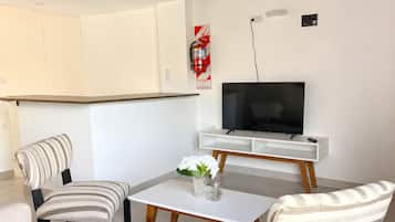 Una sala de estar moderna con un televisor de pantalla plana, dos sillones a rayas, una mesa de centro blanca y un pequeño jarrón con flores.