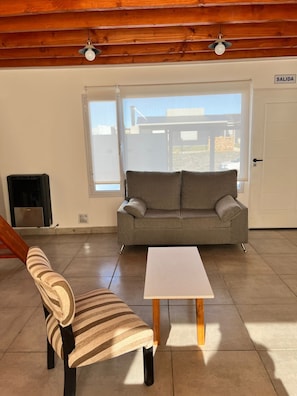 Un salón espacioso con un sofá gris, un sillón a rayas y una mesa de centro blanca. Hay una puerta corrediza de cristal que da al exterior, y un letrero que dice “SALIDA”. 