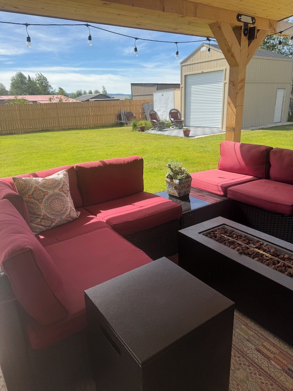 A covered outdoor seating area with red sofas and a small table, a fire feature, and a view of a backyard with a lawn and a building.