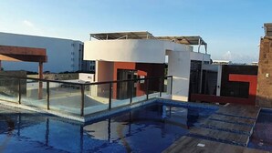 A rooftop pool area with a glass enclosure, wooden deck, and modern buildings in the background. - Walk to the beach from your luxury living apartment (Playa del Carmen)
