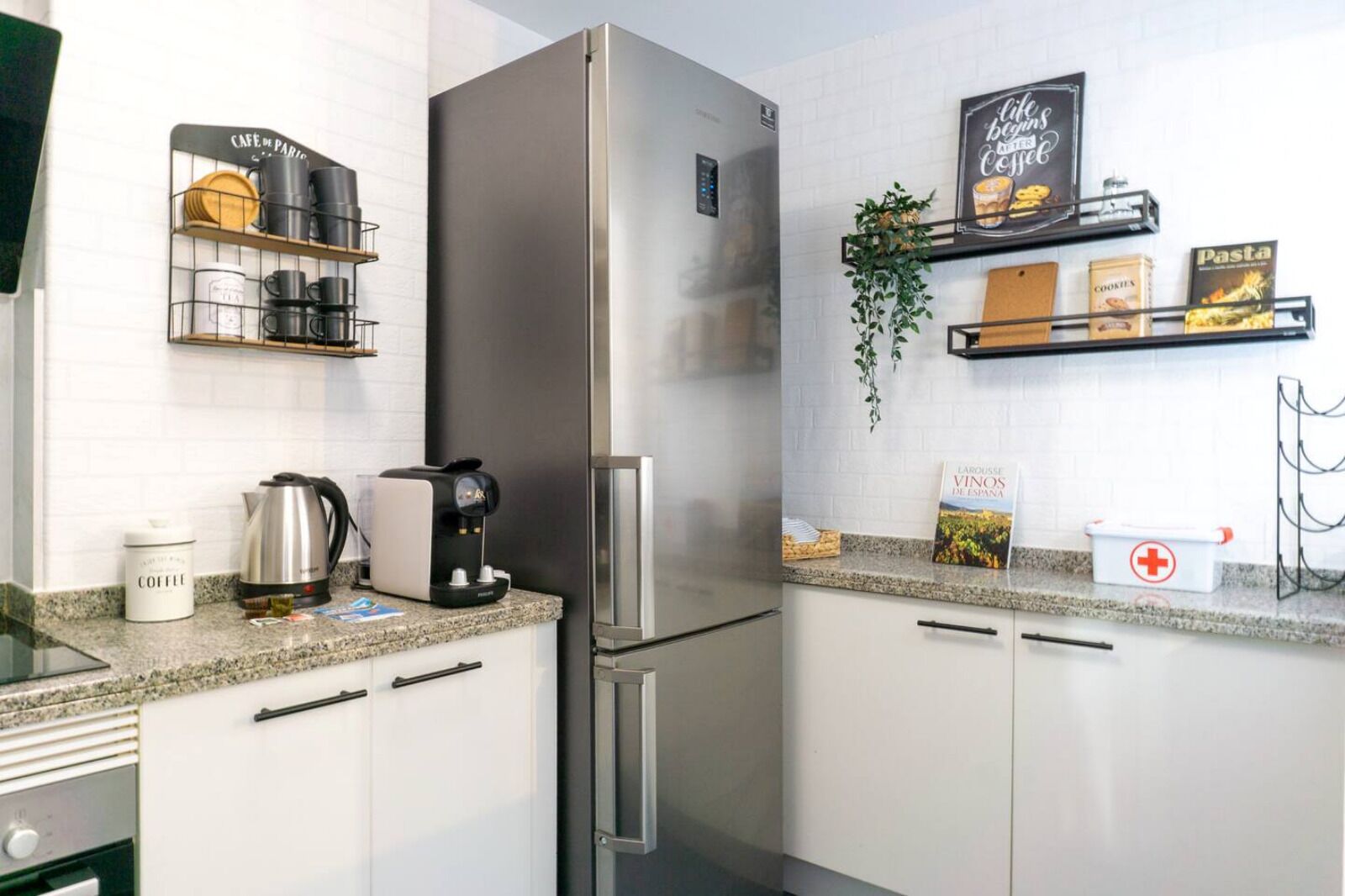 Eine moderne Küche mit einem Edelstahl-Kühlschrank, einer Kaffeemaschine und einem Kaffeebehälter. Es gibt Regale mit Küchenutensilien und ein Buch mit dem Titel 'Pasta'.