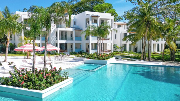 A modern resort with a large swimming pool, lounge areas, and a multi-story building with balconies.