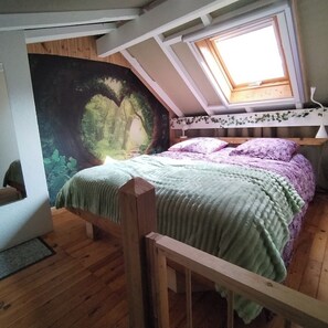 Ein Schlafzimmer mit schrägverdeckter Decke, einem Bett mit einer grün-lila Bettdecke, einem Wandgemälde in Herzform und einem Oberlicht.