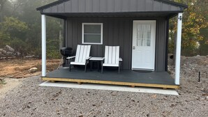 A small, dark-colored cabin with a porch, two Adirondack chairs, and a grill. - Delightful Cabin #4 close to Warrensburg, Knob Knoster, and Clinton. (Warrensburg)