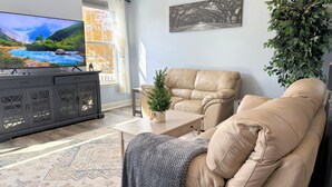 Ein Wohnzimmer mit einem beigen Sofa, einem Fernseher auf einem Schrank, einem kleinen Weihnachtsbaum und einem großen Fenster mit Vorhängen.