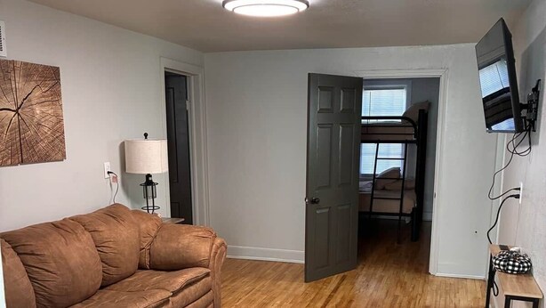 Un salon avec un canapé marron, un tapis à motifs, un plancher en bois, un luminaire au plafond, un téléviseur fixé au mur et une porte donnant sur une autre pièce.
