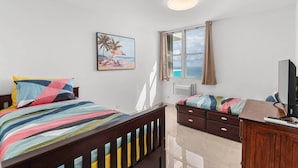A bedroom with a bed, a desk with a computer, a window with curtains, a radiator, and a painting of a beach scene.
