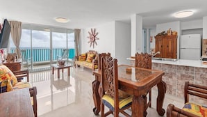 A spacious living area with a balcony, a dining table with chairs, and a kitchen area with wooden cabinets and a refrigerator.
