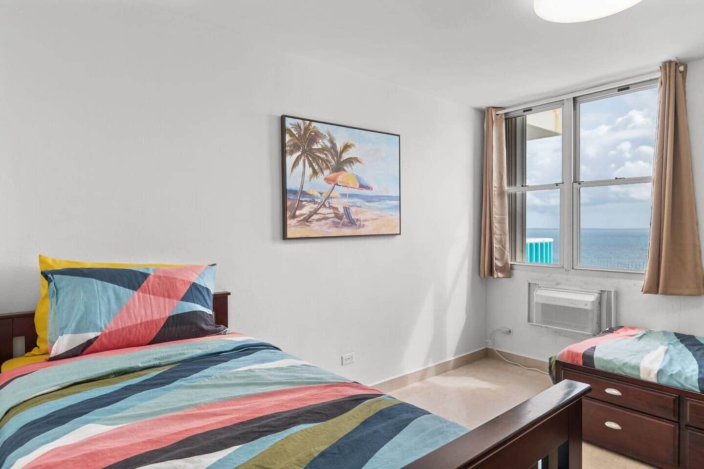 Ein Schlafzimmer mit Bett, Nachttisch, einem Fenster mit Meerblick und einem Bild an der Wand.