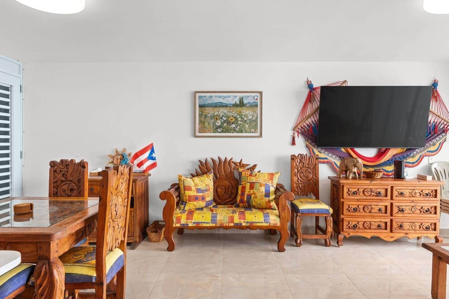 A living room with a wooden sofa, a coffee table, a TV mounted on the wall, and a colorful woven chair.