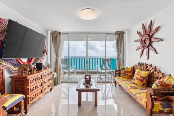 Un salón amplio con televisor grande, muebles de madera, un sofá colorido y un balcón con vistas al océano.
