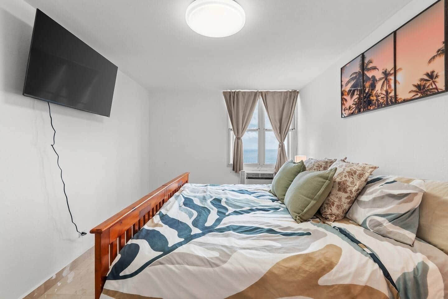 Ein Schlafzimmer mit einem Bett, einem an der Wand montierten Fernseher, einem Fenster mit Vorhängen und einem Gemälde.