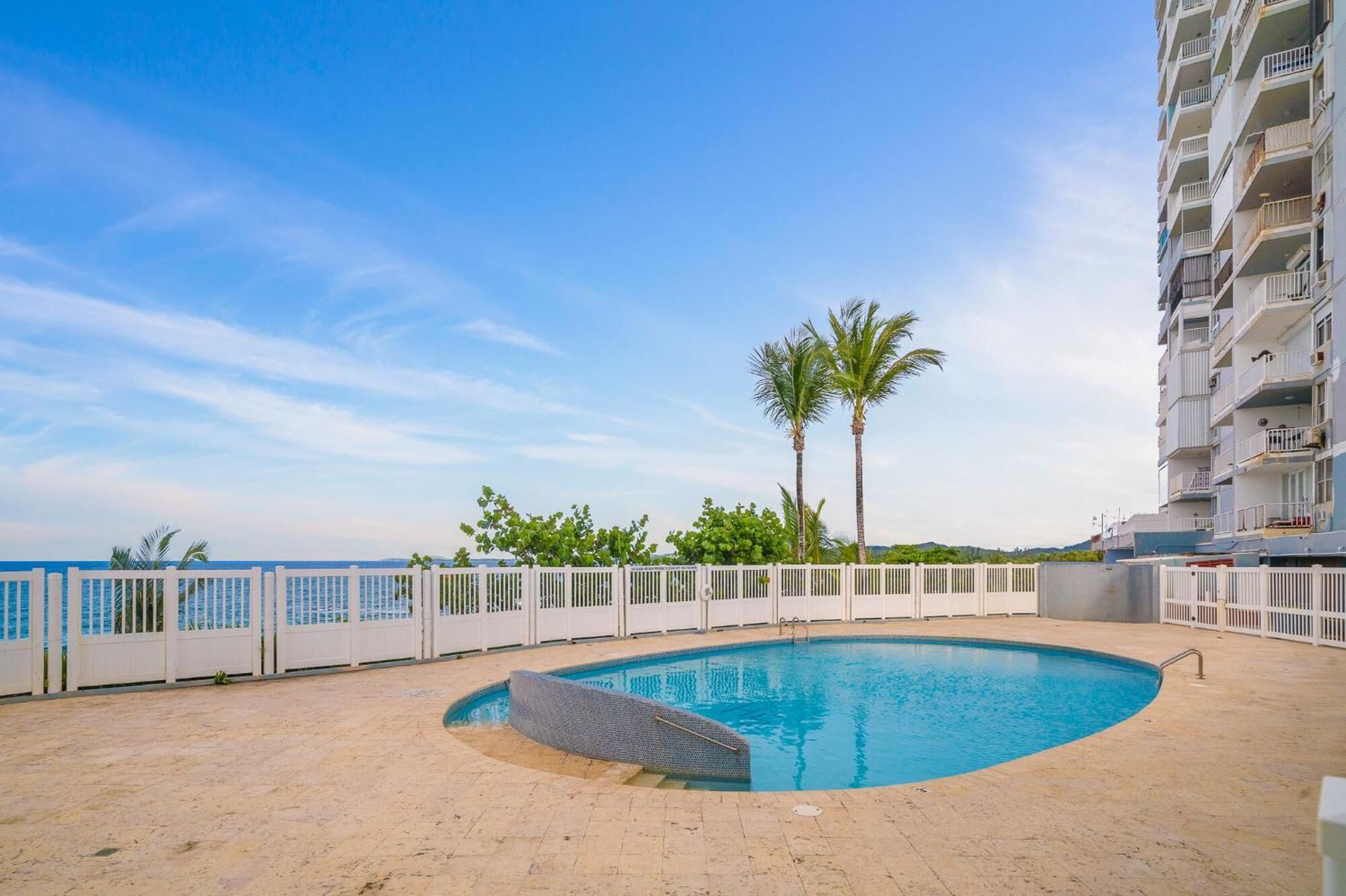 A swimming pool with a white fence, palm trees, and a building with balconies.