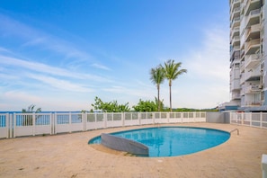 A swimming pool with a white fence, palm trees, and a building with balconies.