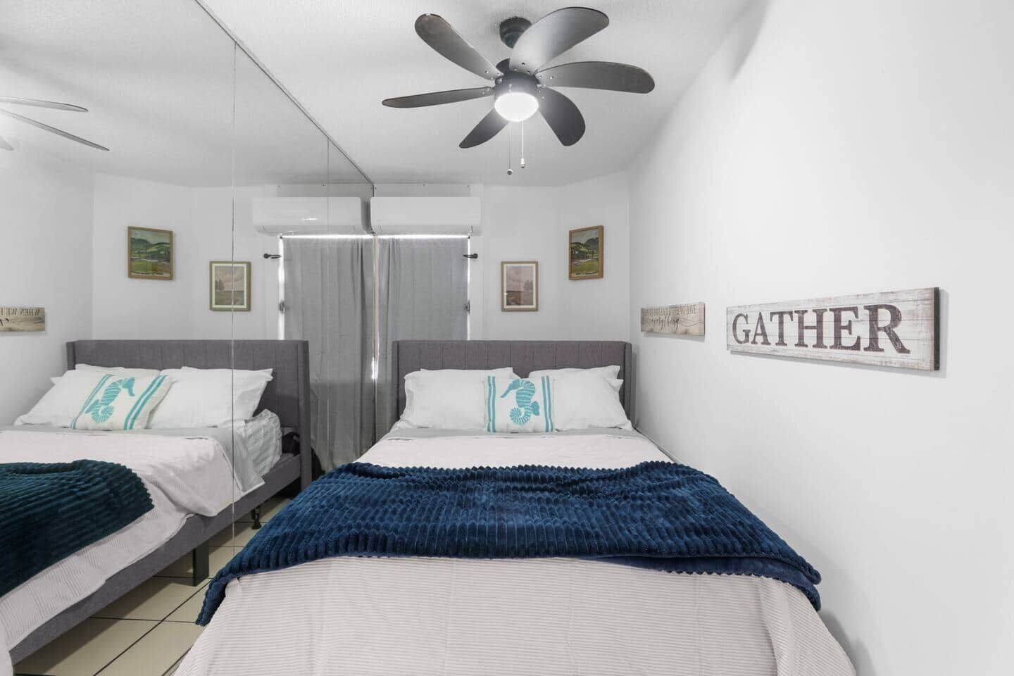 Una camera d'albergo con due letti, una ventola a soffitto e un cartello con la scritta 'GATHER'.