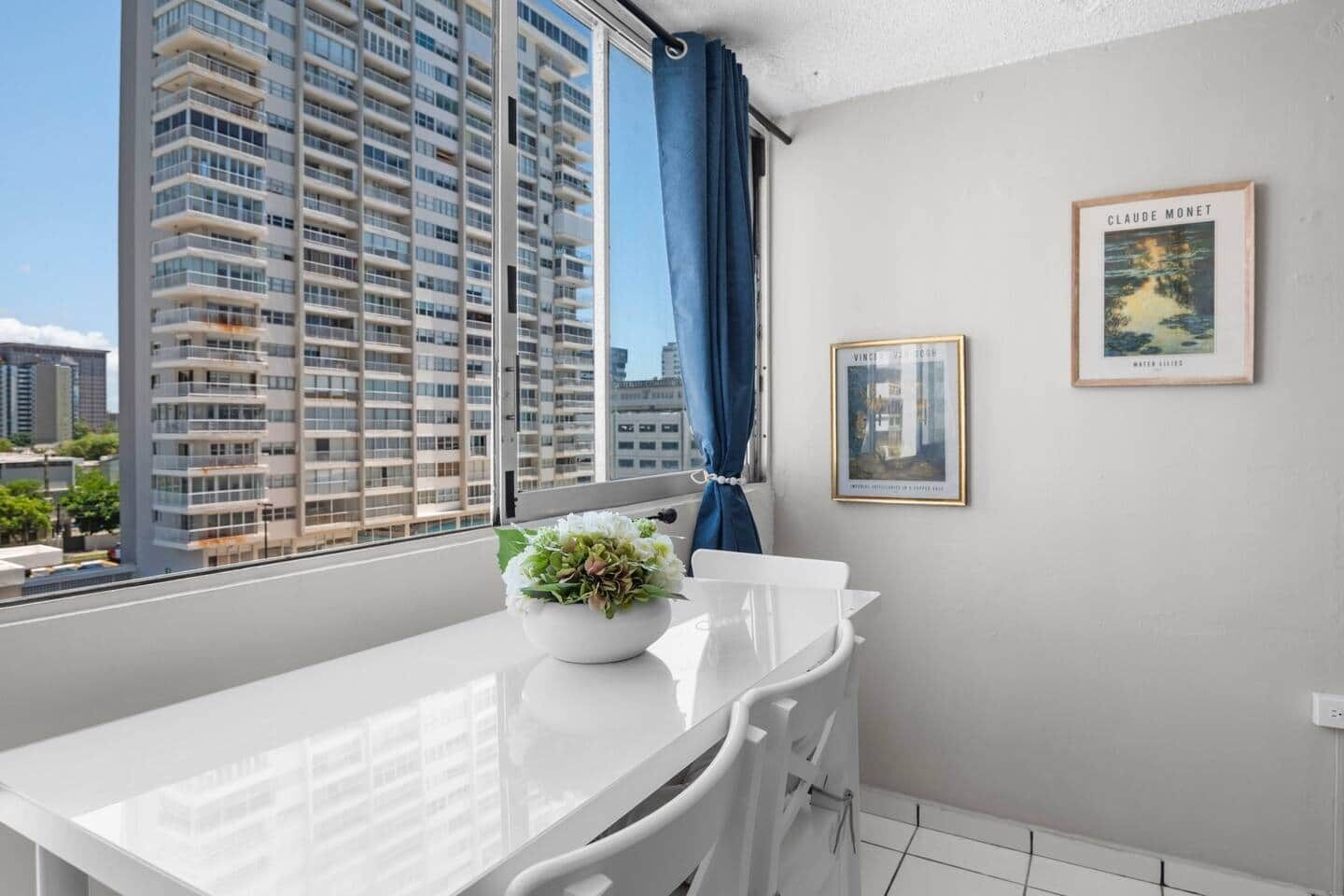A modern apartment with a white dining table, a vase of flowers, and a view of high-rise buildings.