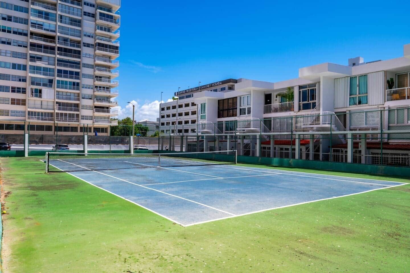 An outdoor tennis court with a net, surrounded by modern buildings, including a hospital.