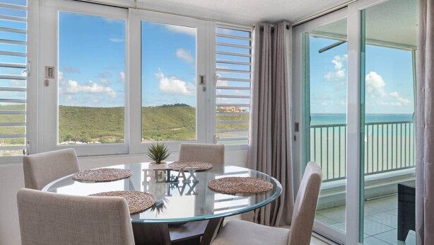 Une table ronde en verre, accompagnée de quatre chaises, d’une petite plante en pot et d’un objet décoratif portant l’inscription « RELAX », le tout placé sur un balcon offrant une vue sur la mer et les collines.