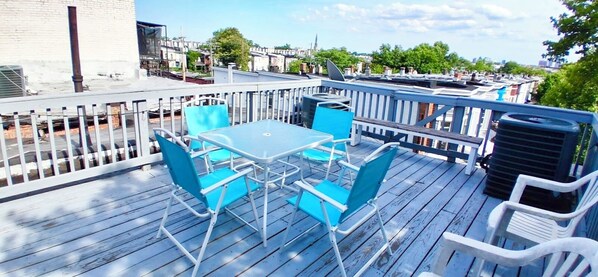 Une terrasse sur le toit, avec une table et des chaises, une rambarde et une vue sur les bâtiments et les arbres.