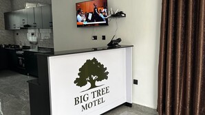 A reception desk with a TV screen displaying a band, a logo for 'Big Tree Motel', and a kitchen area in the background. - BIG TREE MOTEL (Ruhengeri)