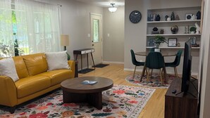 A living room with a yellow sofa, a round coffee table, and a floral rug. There is a dining area with a small table and chairs, and a TV stand with a television. - Par-fect Escape on Lake Windsor Golf Course (Windsor)