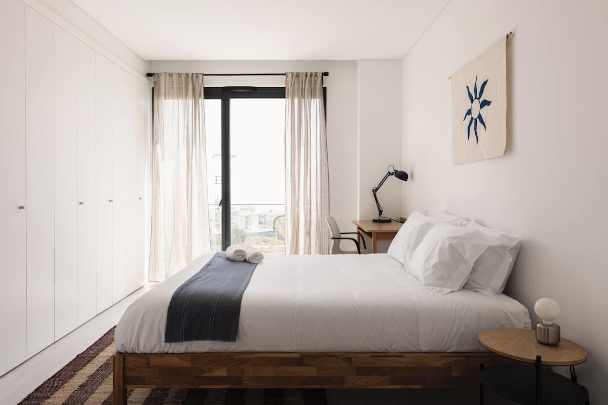 Ein modernes Hotelzimmer mit einem großen Bett, einem Schreibtisch, einem Stuhl und Blick auf die Stadt durch das Fenster.