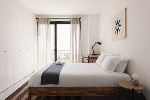 Un dormitorio moderno con una cama de madera, ropa de cama blanca, un escritorio con lámpara y vistas a través de un amplio ventanal con cortinas.