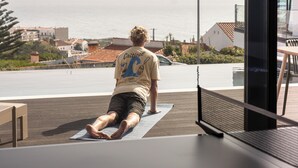 Une personne fait du yoga sur un tapis près d’une grande fenêtre donnant sur un plan d’eau et des bâtiments.