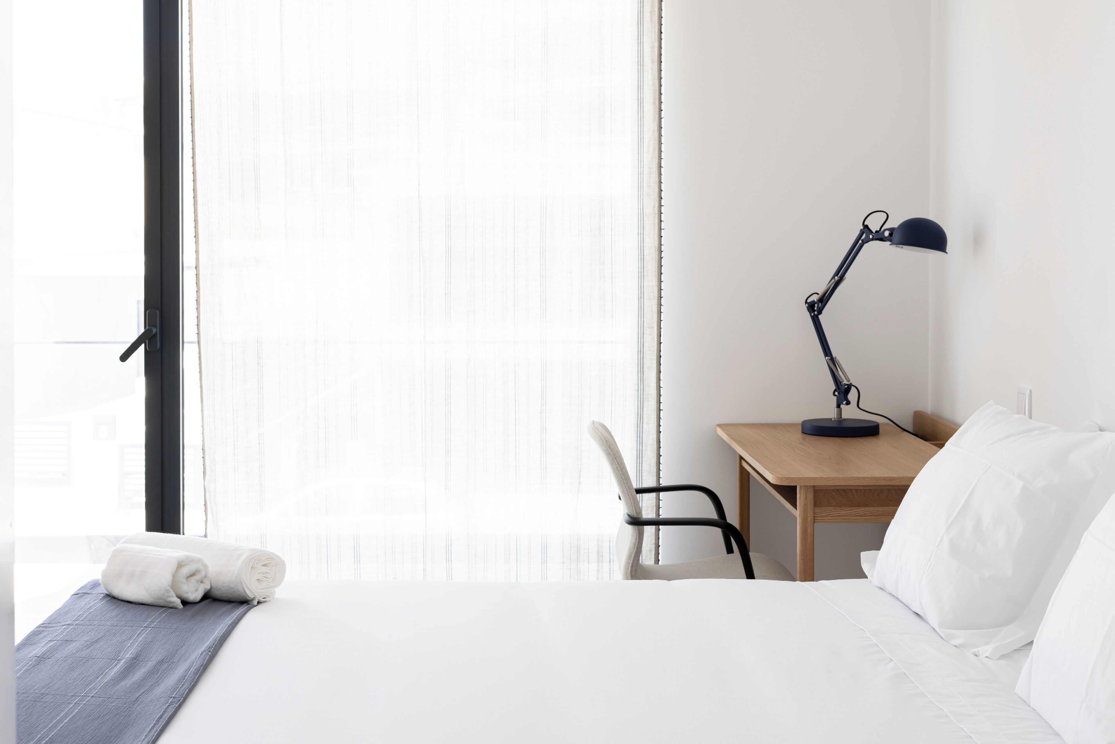 Una camera d'albergo con un letto, una scrivania con una lampada, una sedia e una finestra con tende leggere.