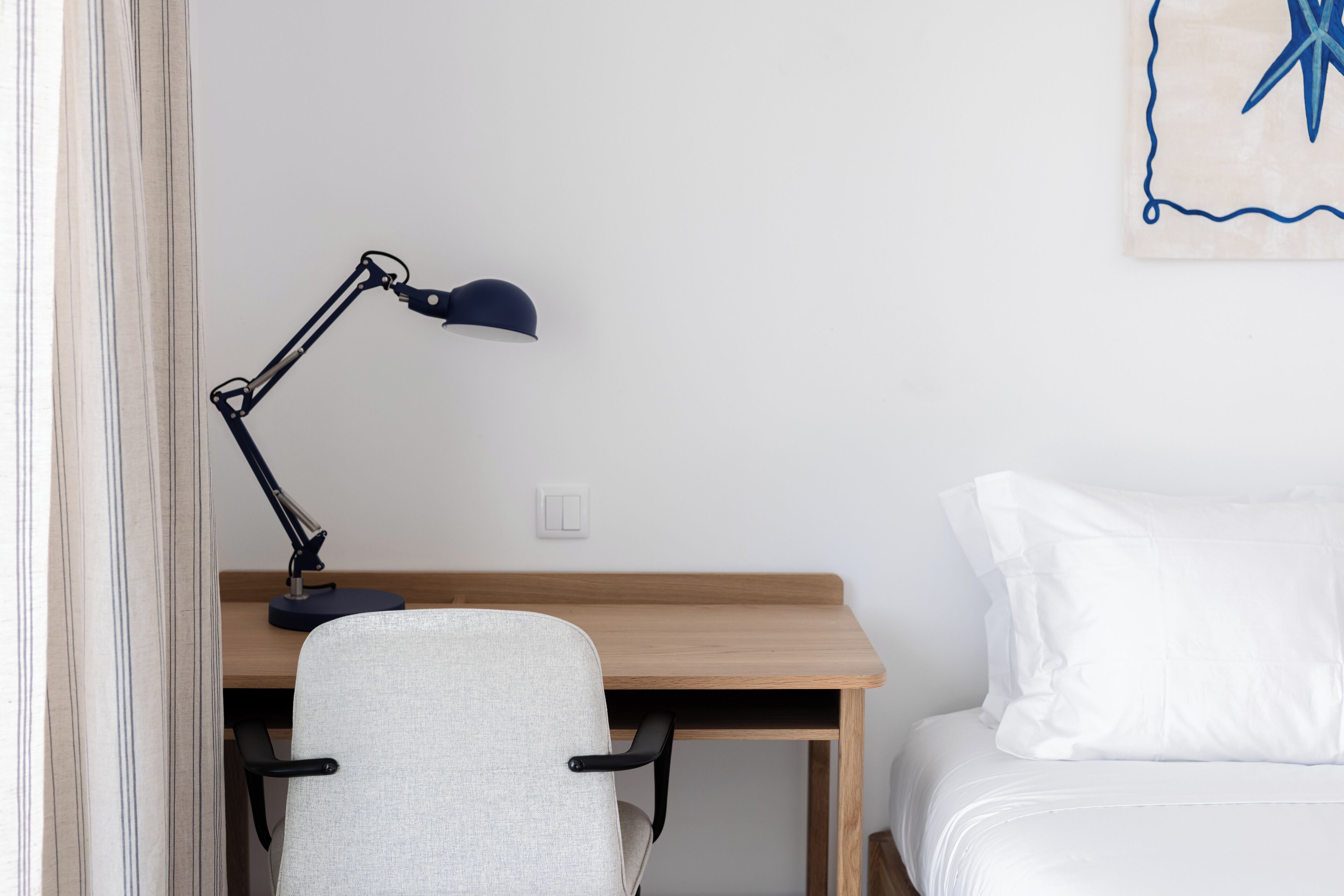 A bedroom with a wooden desk, a chair, a lamp, and a bed with white bedding.