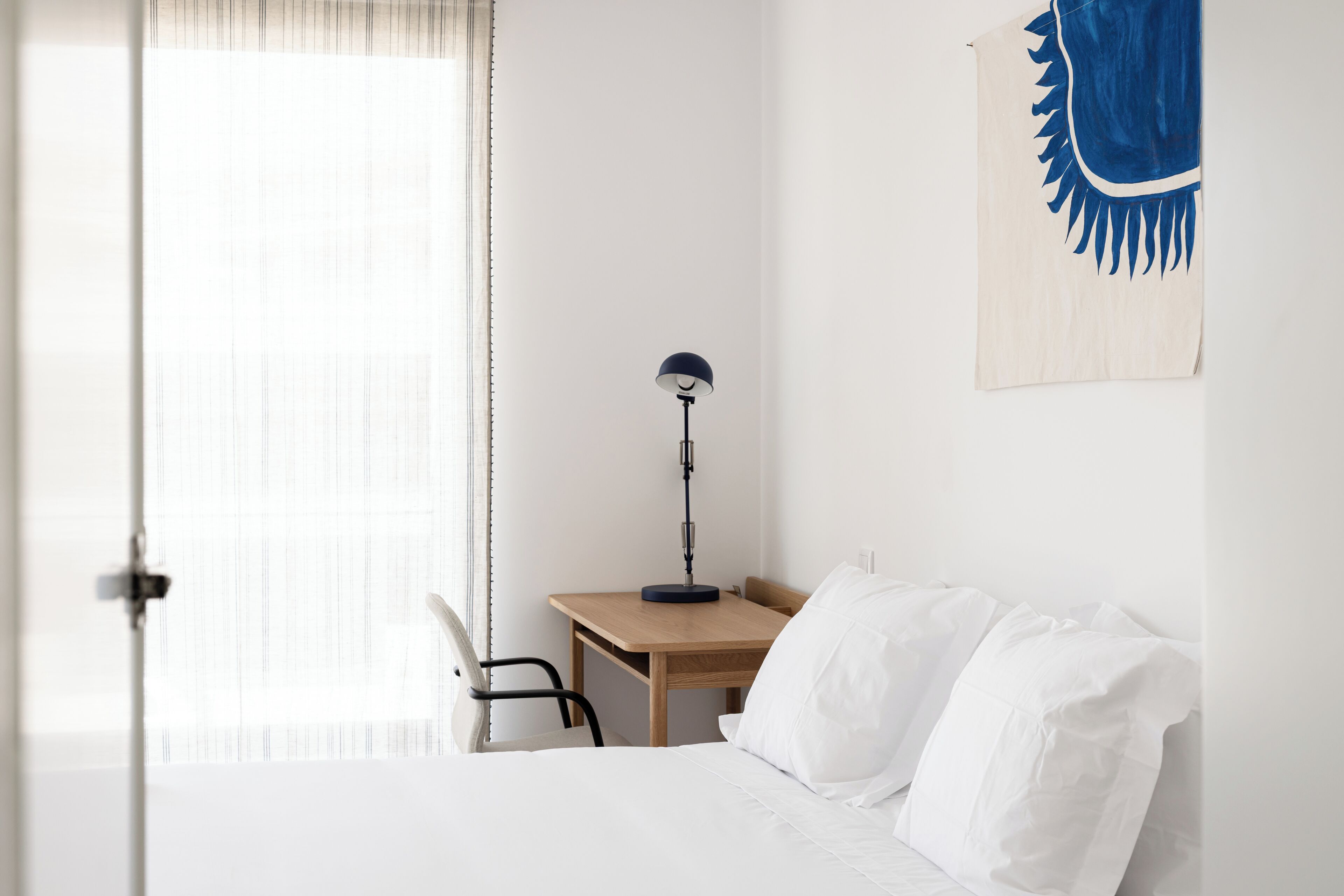 A hotel room with a bed, a desk, a chair, a lamp, and a window with curtains.