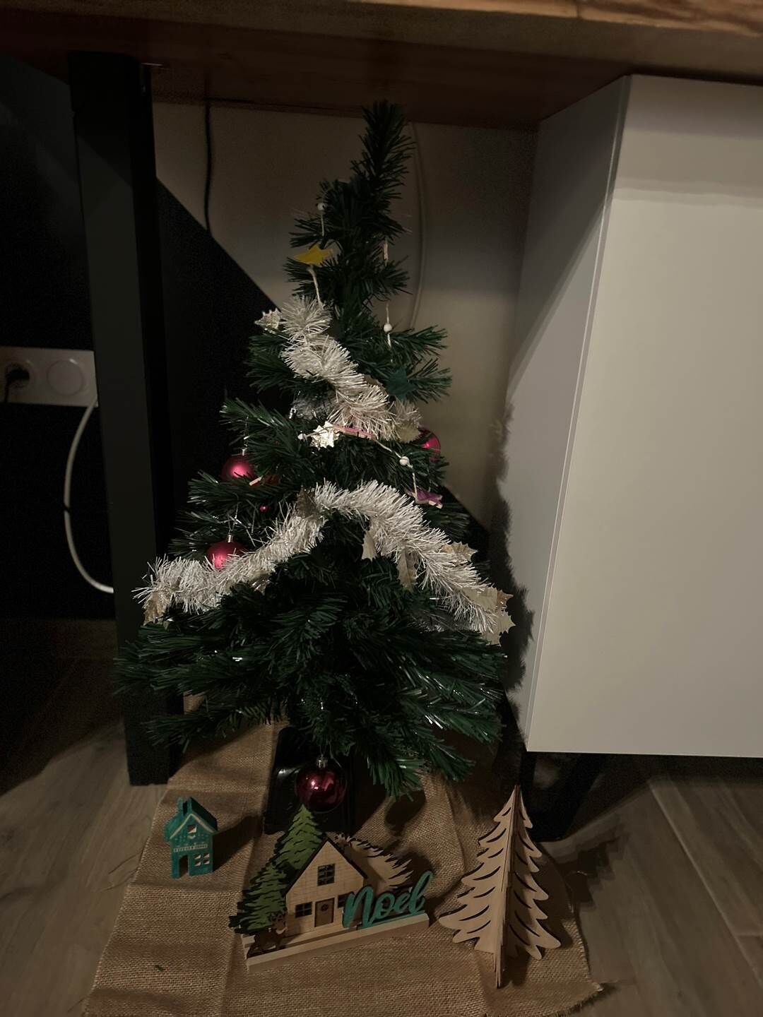 Een kleine kerstboom met decoraties, een houten huisje en het woord 'Noel' op een linnen zak.