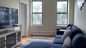A living room with a TV, a sofa, and a window. - NYC Skyline Apartment (Weehawken Township)