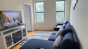 A modern living room with a sectional sofa, a flat-screen TV, and a large window providing natural light. - NYC Skyline Apartment (Weehawken Township)
