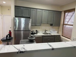 Una cocina con gabinetes grises, un refrigerador de acero inoxidable, encimera de mármol y una ventana con persianas.
