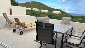 A terrace with a table and chairs, a wheelchair, and a view of buildings on a hill. - Changes in Latitude 18.3 spacious Mahogany Run condo with ocean views. (St Thomas)