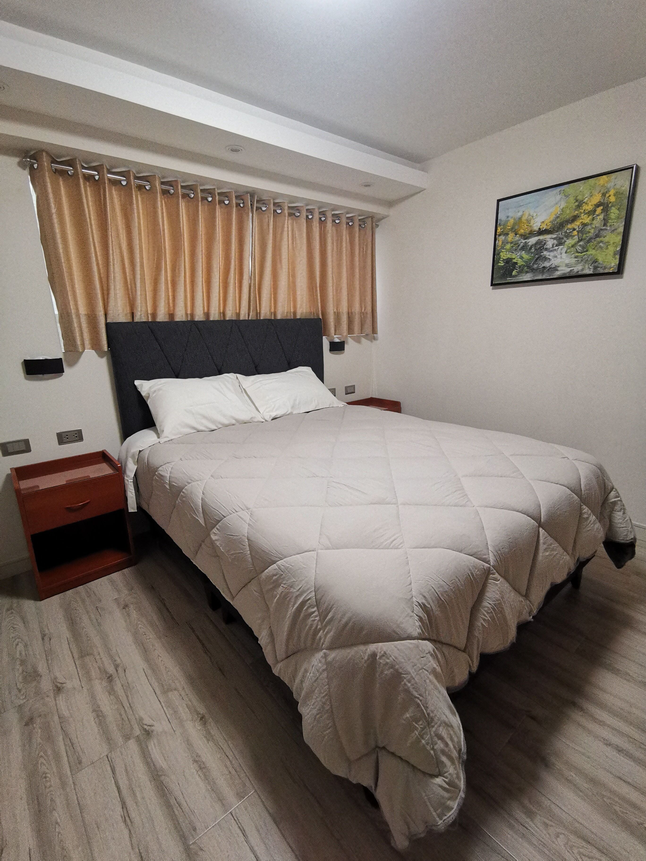 A neatly made bed with a quilted cover, a wooden nightstand with a drawer, a wall-mounted power outlet, and a painting of a landscape on the wall.