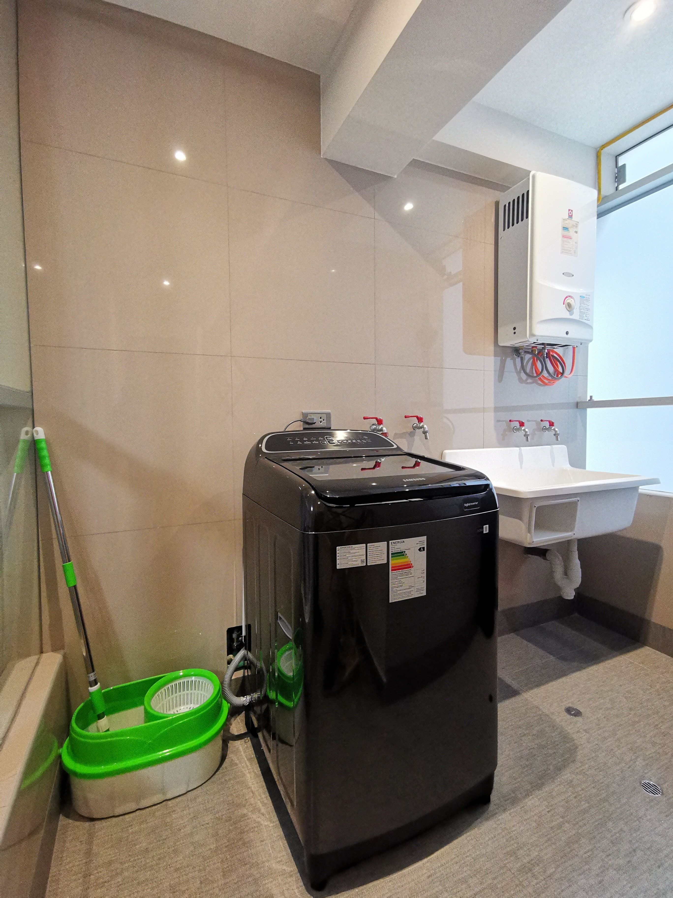 A bathroom with a black water heater, a green and white mop bucket, a white sink, and a mirror.
