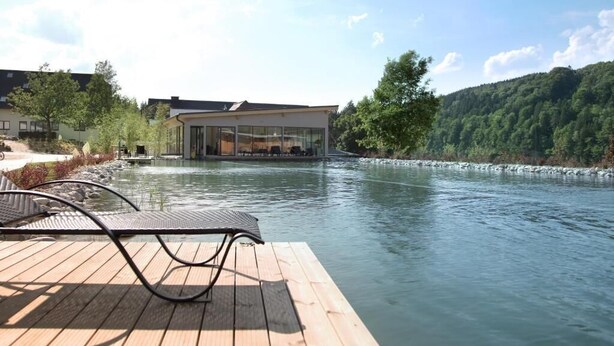 Blick auf einen Fluss mit einer Holzterrasse und einem Glasgebäude.