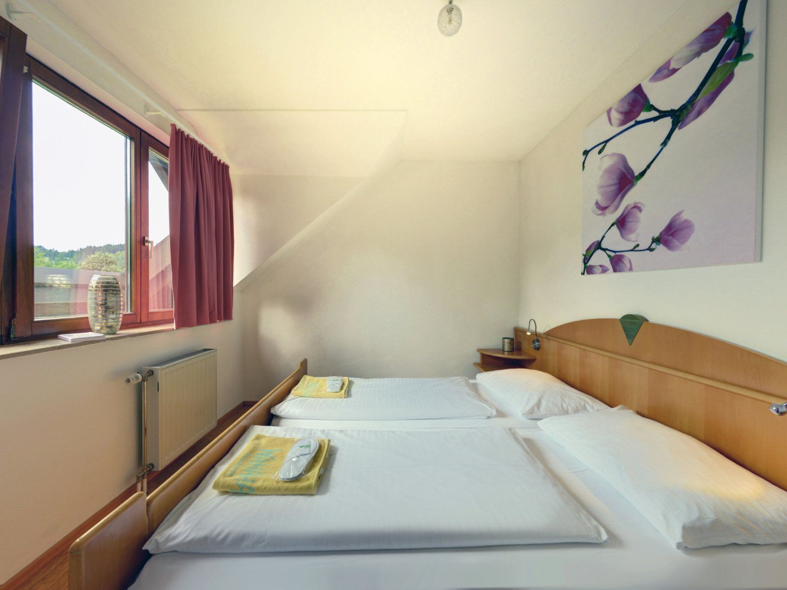 Une chambre d’hôtel avec deux lits, une fenêtre avec des rideaux, un radiateur, une table de chevet avec une lampe et un tableau au mur.