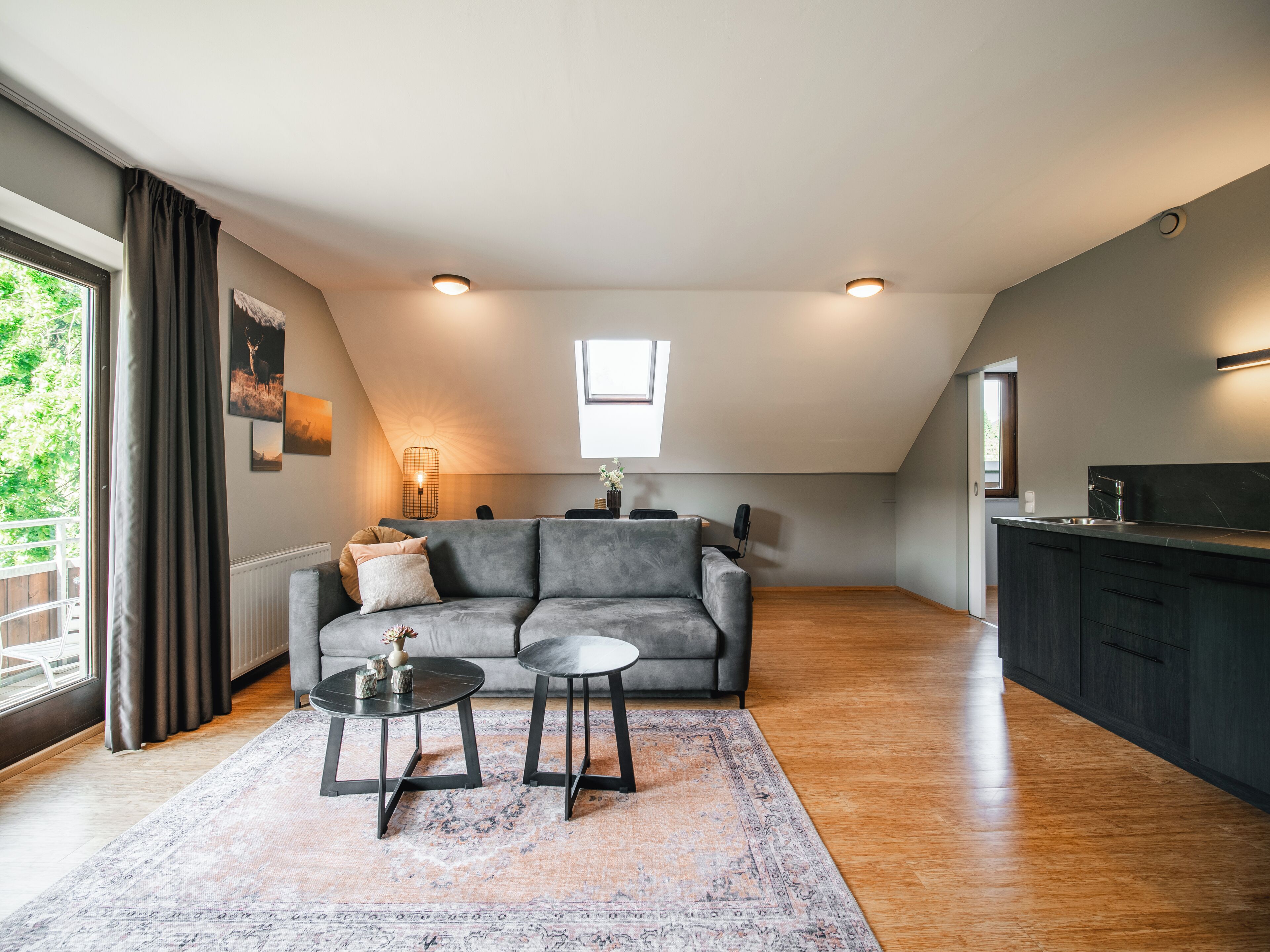 A modern living room with a grey sofa, two small tables, a patterned rug, and a kitchen area with dark cabinetry.