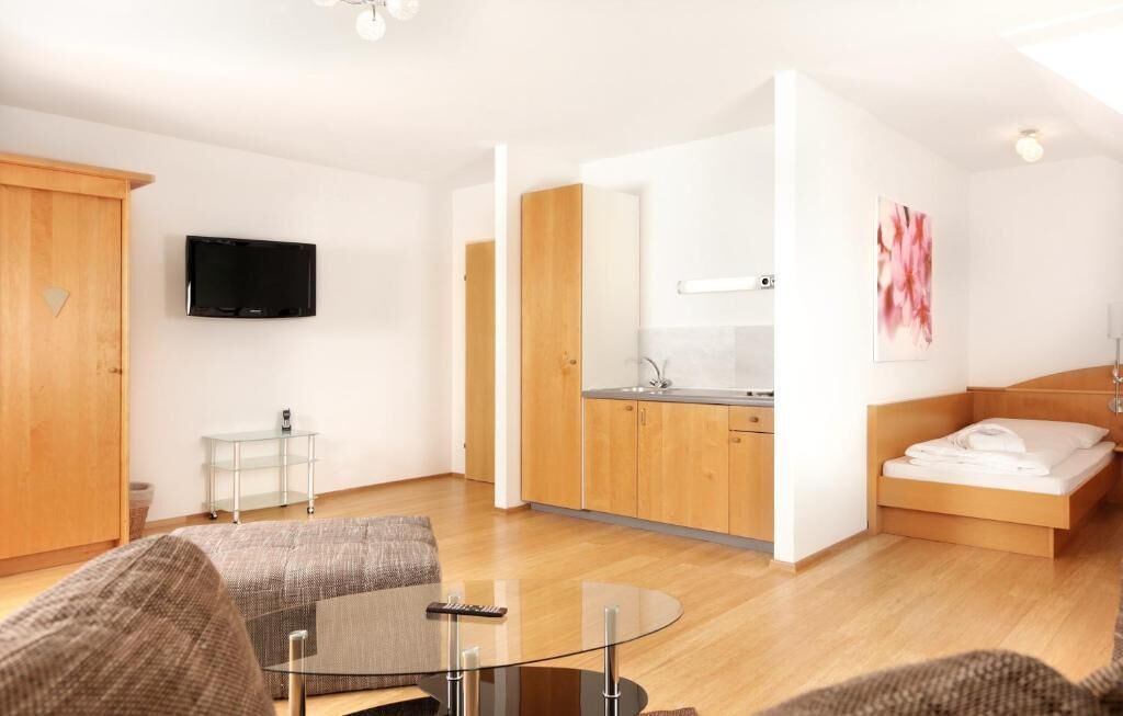 A modern hotel room with a wooden floor, a glass table, a sofa, a bed, a TV, and a kitchenette.