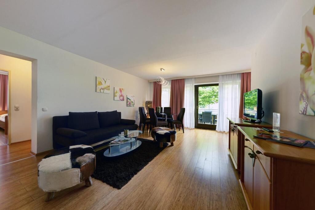 Una sala de estar moderna con sofá, mesa de centro y reposapiés. Hay una zona de comedor con sillas y una mesa. Un televisor está montado en la pared.