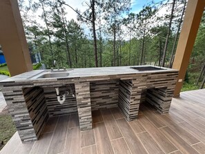 An outdoor kitchen with a sink and a grill, surrounded by a forested area. - Casa Di Valle (Valle de Ángeles)